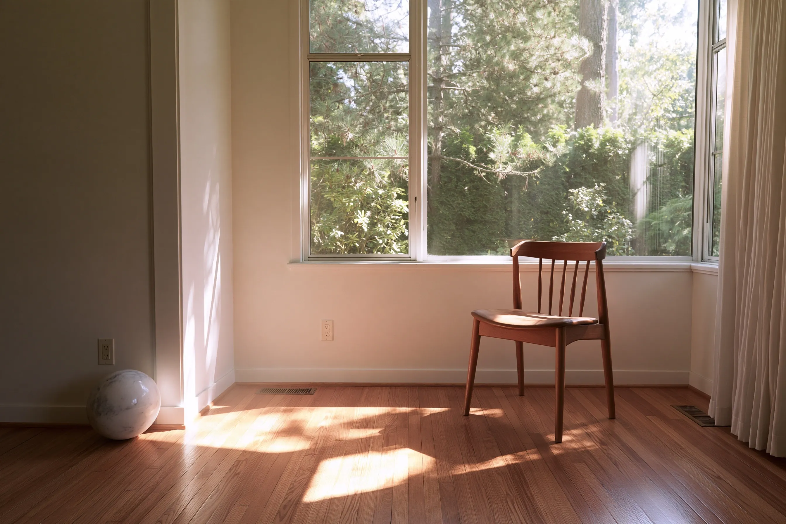 Empty living room with a hardwood floor, a wooden chair by a large window onto trees and a small ball resting against the wall in the corner